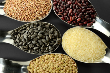 Rice, buckwheat, seeds, lentils, beans in spoons on a dark background.