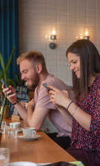 two friends sitting together in a cafe, using smart-phones. sitting together, yet each is using at their phone.
