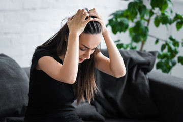 frustrated young woman suffering from depression while sitting on couch and holding hands on head