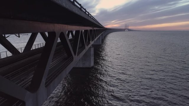 Drone shot flying up next to &Oslash;resund Bridge at dusk. Aerial view over sea and railway bridge, train tracks and driving cars at sunset. Connection between Malm&ouml;, Sweden and Copenhagen, Denmark
