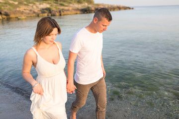 Outdoor shot of romantic young couple walking along the sea shore holding hands. Young man and woman walking on the beach together at sunset, body closeup