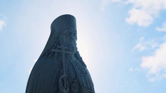 Close-up view of bronze or stone statue of priest with cross in his hand in sunny day against blue cloudy sky. Stock footage. Religion and faith concept