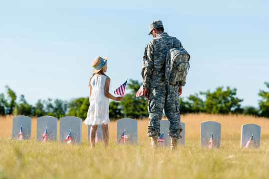 child in straw hat and man in military uniform standing near headstones and holding american flags in graveyard