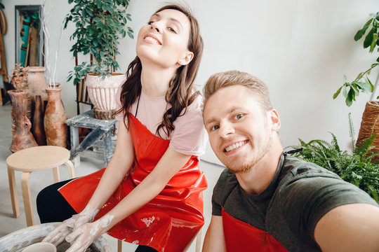 Concept loving couple at masterclass of artists. Happy man makes selfie photo on background of girl behind a potter wheel, dishes from clay