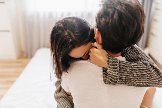 Brown-haired woman in woolen cardigan hiding face on husband's shoulder. Indoor photo of shy girl in gray soft sweater hugging with boyfriend in light bedroom.