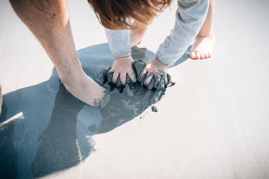 From above barefoot active kid and parent having fun with wet sand touching by hands in sunny day