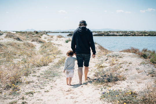 Back view of young man in dark clothes holding hands with kid and walking around sandy shore with dry greenery in sunny day