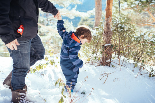 Young man in dark warm clothes holding hands with kid in striped overalls and walking in snow in sunny day