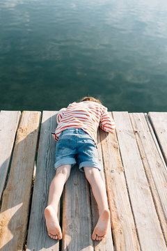 From above of chilling kid lying with face down on wooden pier in sunshine with peaceful water below