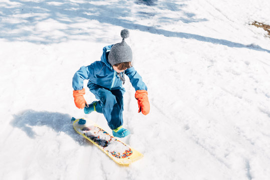 Boy in winter clothes riding small board while sliding down snowy mountain in sunlight