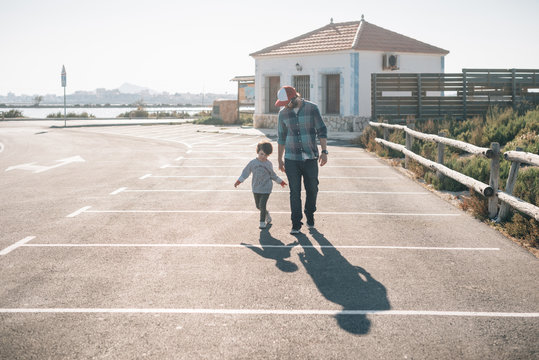 Adult man with playful son in casual clothes walking on parking of street in sunshine