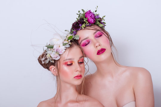 Two fashion beauty models sister girls with professional bright make-up, a hoop of fresh spring flowers on their heads, with red lilac lipstick are posing against a gray uniform background. Clean skin