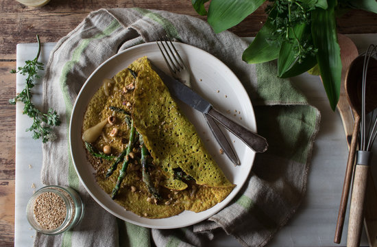 Top view of white plate with oat crepe with asparagus and tahini paste served on rustic board with greens