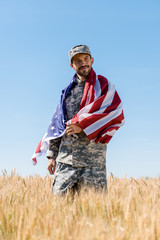 selective focus of happy soldier in cap and uniform holding american flag in field