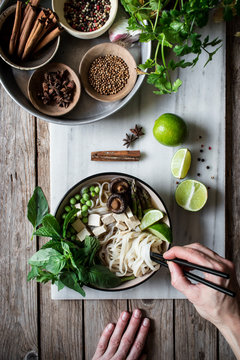 From above person hands with arranged bowls with dry spices and served Pho soup with noodles on marble board