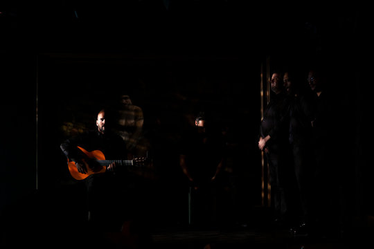 Hispanic men playing percussion and acoustic guitar during flamenco performance on dark stage