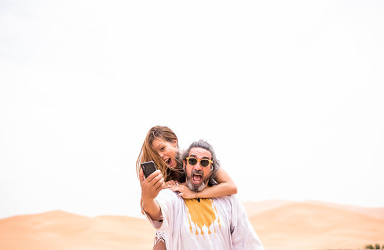 Middle-aged man with woman piggyback taking selfie expressively on terrace against sandy desert, Morocco