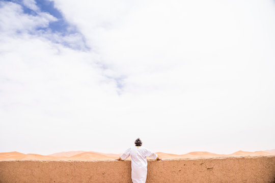 Back view of anonymous adult man in long white clothes leaning on a wall looking away against endless sandy desert, Morocco