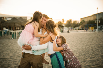 Adult loving man and woman kissing while holding an embracing with daughters standing together on beach in back lit