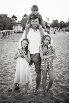 Adult man with laughing boy on shoulders standing with beautiful little girls on beach looking at camera, black and white photo