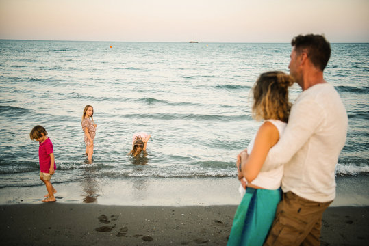 Side view of embracing man and woman looking at playful boy and girls in water on coastline