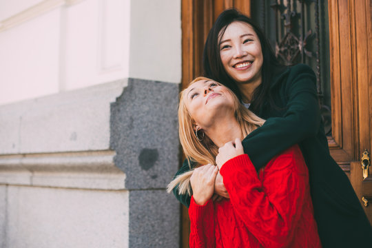 Happy Asian female smiling and looking at each other while standing against ornamental door of aged building and embracing Caucasian friend