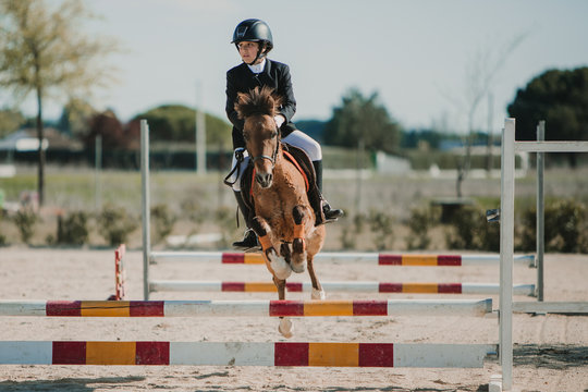 Teenage jockey on horse leaping over horizontal wooden bars while riding on racetrack