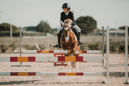 Teenage jockey on horse leaping over horizontal wooden bars while riding on racetrack