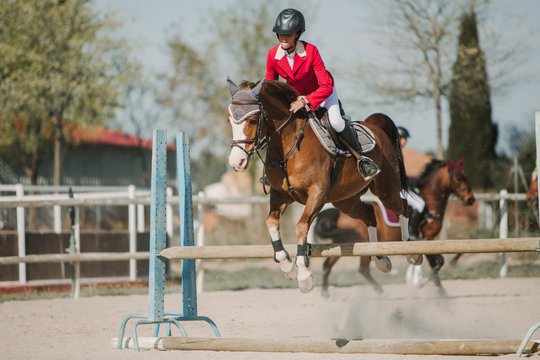 Teenage jockey on horse leaping over horizontal wooden bars while riding on racetrack