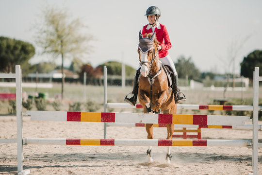 Teenage jockey on horse leaping over horizontal wooden bars while riding on racetrack