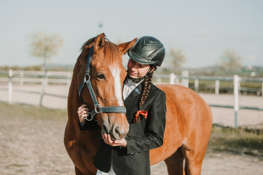 Side view of young teen woman in jockey helmet and jacket caressing horse standing together outdoors