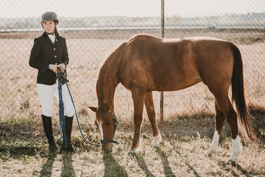 Teenage girl in jockey outfit standing with chestnut horse pasturing near fence of ranch