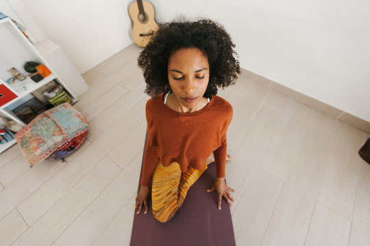 From above face of African American attractive young woman sitting in yoga pose with closed eyes on mat in light room