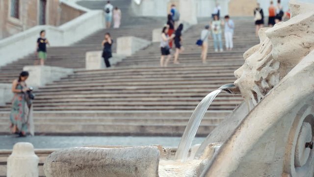 Roman marble fountain with interesting monument streaming water on the background of ancient famous Spanish Steps. Tourists visiting place in the morning and enjoying surrounding landmark sitting on