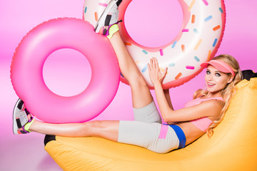 Obraz na płótnie Canvas attractive happy girl on bean bag chair with inflatable swim rings on pink, doll concept