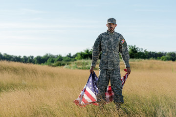 handsome soldier in camouflage uniform holding american flag in golden field