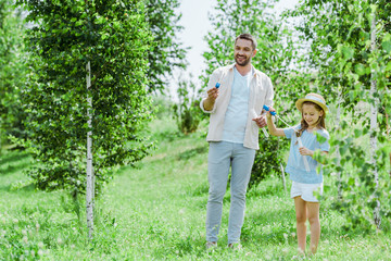 selective focus of cheerful father and daughter holding bubble wands while standing near trees
