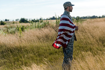 Side view of soldier in camouflage uniform holding American flag in golden field