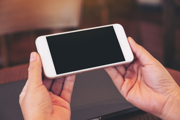 Mockup image of woman's hands holding black mobile phone with blank white screen with laptop computer on wooden table