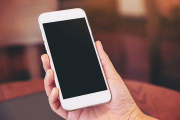 Mockup image of woman's hands holding black mobile phone with blank white screen with laptop computer on wooden table