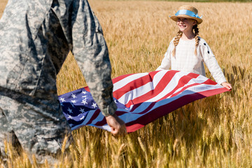 cropped view of man in military uniform holding american flag with cute daughter in field