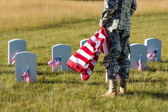 Cropped View Of Man In Military Uniform Holding American Flag While Standing In Graveyard