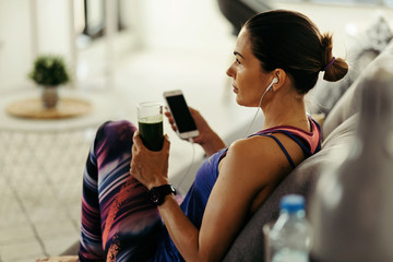 Pensive sportswoman listening music and drinking smoothie on the sofa.