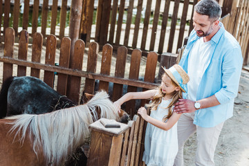 cheerful kid touching pony while standing near father in zoo