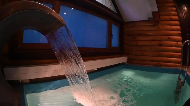 water pours from an artificial waterfall into a pool with a ladder, a log sauna, a wide angle slow motion