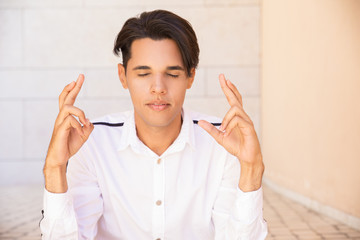 Serene Latin man keeping fingers in mudra gesture and meditating. Handsome young man wearing shirt,...
