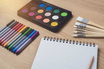 Naklejka premium Notepad or notebook with Many colorful pens, paintbrush and Watercolor palette on brown wood table.using for Arts and Education background