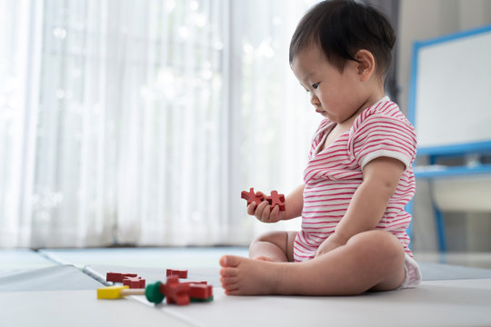 Asian Cute Baby Sitting And Playing A Small Toy On Soft Mat At Home. The Kid Holding The Toy And Question About How To Play It. Play And Learn Of Young Child Education Concept. Seen From Child Side.