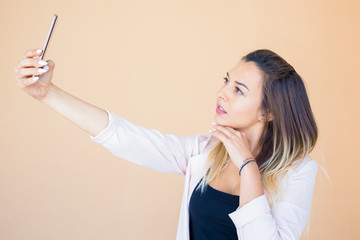 Pensive beautiful lady posing for smartphone camera. Serious young woman taking selfie over beige...