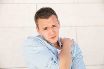 Tensed young man touching shoulder at wall outdoors. Handsome white guy wearing jeans shirt, looking at camera and frowning with building wall in background. Pain concept. Front view.